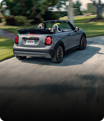 Rear-view of a MINI Cooper S Convertible in Icy Sunshine Blue turning around a bend in the road with a blurred, blue building in the background.