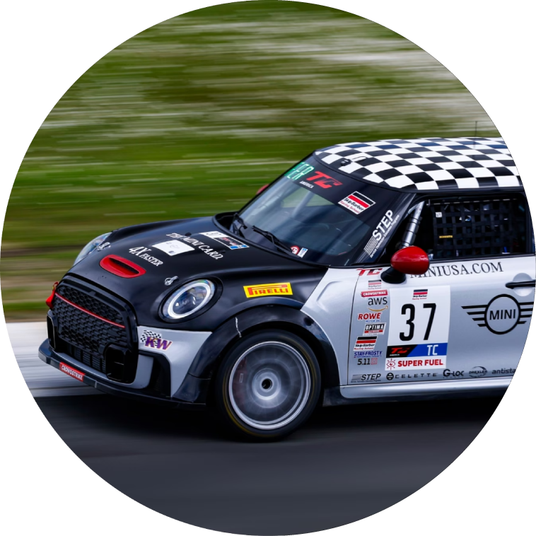 Side view of a MINI race car with a black-and-white checkered roof driving on a pavement surface with its shadow underneath it and surroundings blurred out.