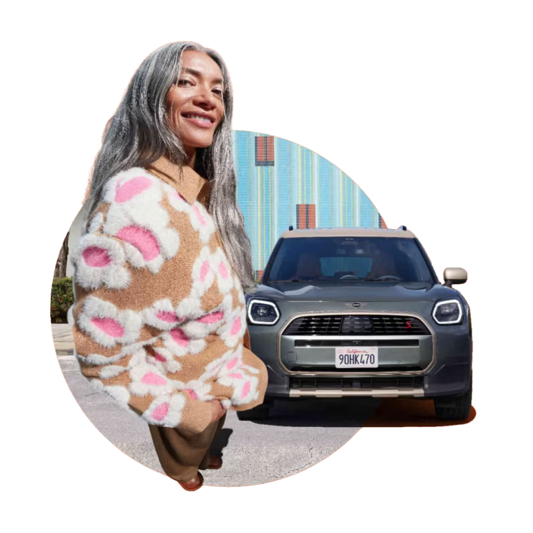 Front view of a MINI Countryman S ALL4 parked on an asphalt surface with its shadow underneath it and a striped wall in the background, surrounded by a middle-aged woman in a fisheye view on the left side of the vehicle.