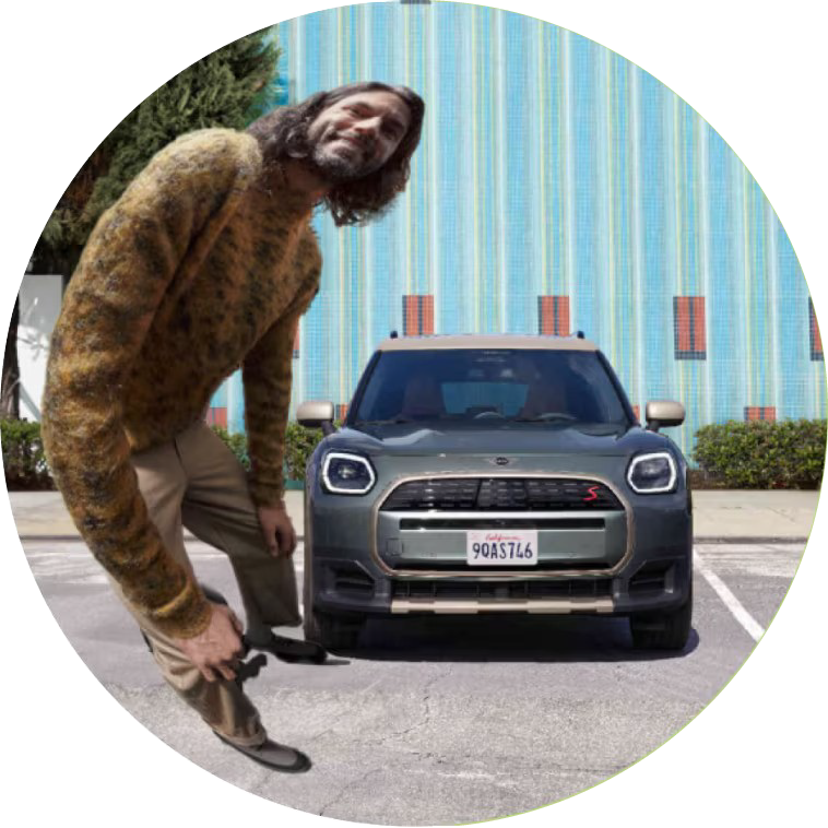 Front view of a MINI Countryman S ALL4 parked in a parking lot in front of a striped wall, with a fisheye view of a man in a brown sweater standing next to it.