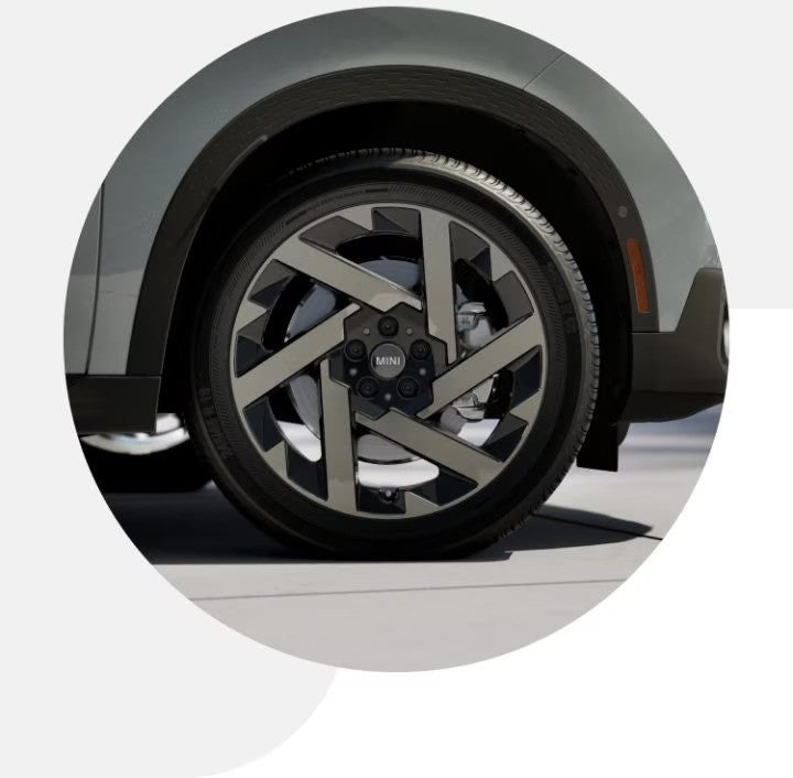 Closeup view of a tire on a MINI Countryman S ALL4, parked on a pavement surface with a shadow underneath the vehicle.