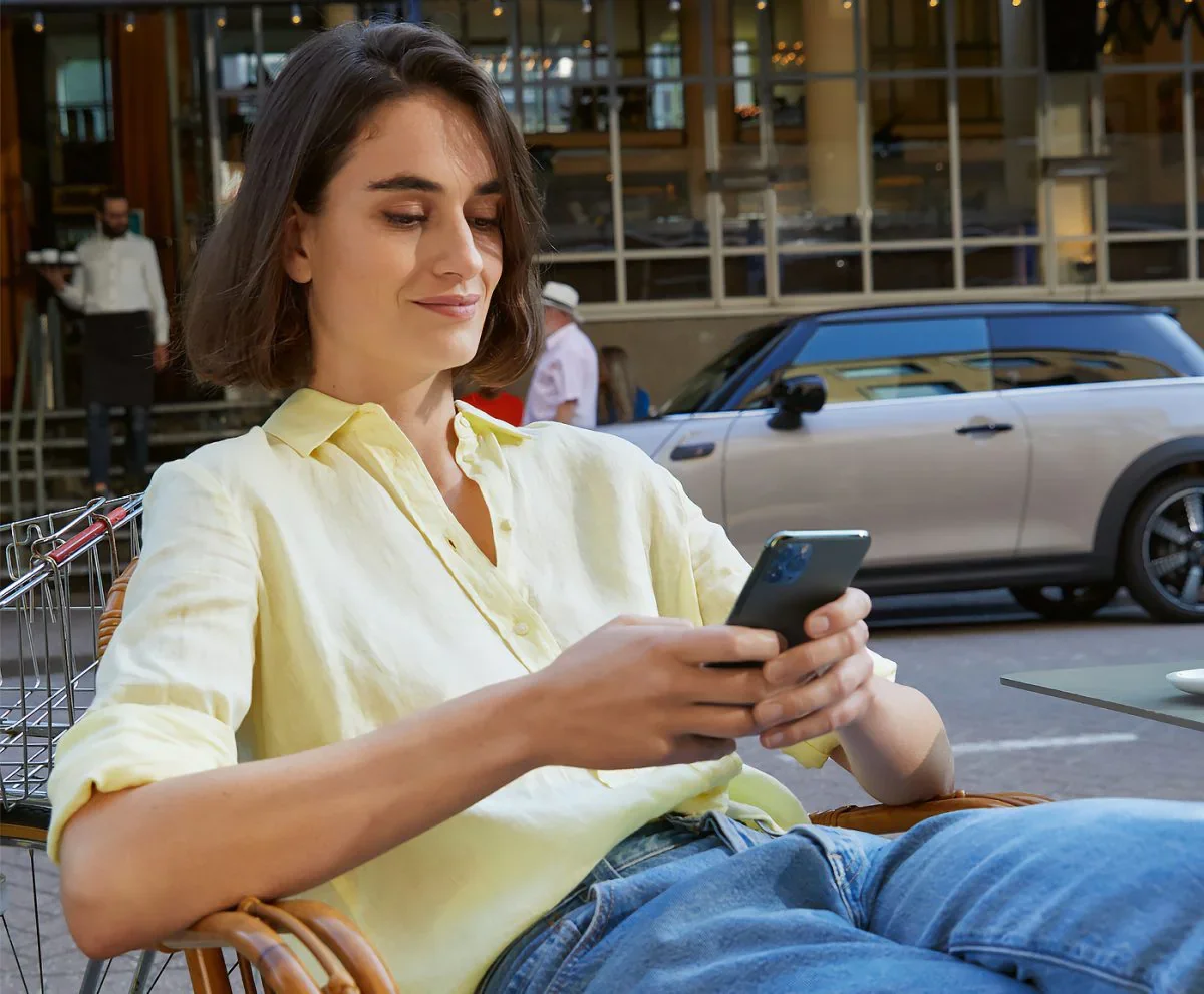 Female wearing yellow collared button-down shirt and jeans, sitting in a chair and holding a smartphone next to a street with a grey MINI vehicle parked in the background. | MINI of Madison in Madison WI